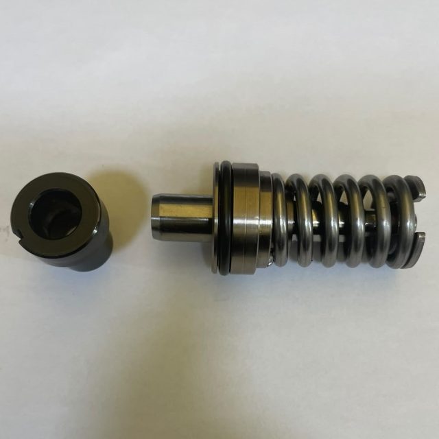 Metal compression damper with a coiled chrome spring and cylindrical plunger, laid beside a round end cap on a white background.